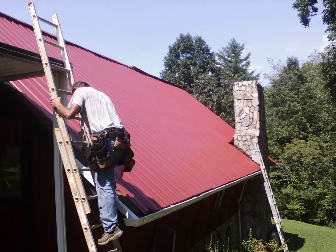 Roofer on a ladder beside a red metal roof and stone chimney — Soffit & Fascia Repair by Roofing Brothers in Pottsville
